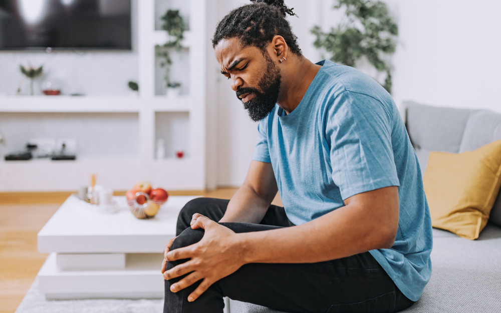 A mid-adult African-American man is sitting in a living room and massaging his painful knee.