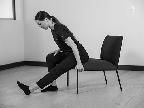 Woman tit closer to the edge of the chair. With one foot flat on the floor, extend the other out straight with the heel on the ground.